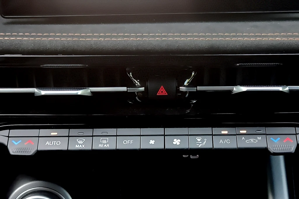 Fizyczny panel sterowania klimatyzacją. Spasowanie przycisków mogłoby być lepsze... Panel sterowania klimatyzacją w nowoczesnym samochodzie, centralnie widoczny przycisk świateł awaryjnych i szereg funkcjonalnych przycisków odpowiadających za regulację temperatury, nawiewu oraz ogrzewania szyb.