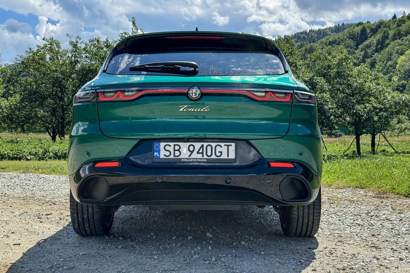 Zielony samochód marki Alfa Romeo Tonale uchwycony od tyłu, zaparkowany na żwirowej drodze, w otoczeniu drzew i wzgórz na tle lekko zachmurzonego nieba.