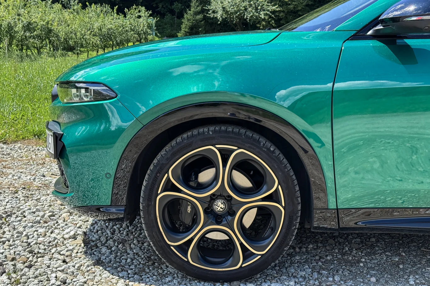 Metaliczny zielony samochód osobowy zaparkowany na żwirowej nawierzchni, widoczny jest szczegółowo przedni lewy błotnik z charakterystycznym aluminiowym kołem o czarnych i złotych akcentach. W tle zielona trawa i drzewa.