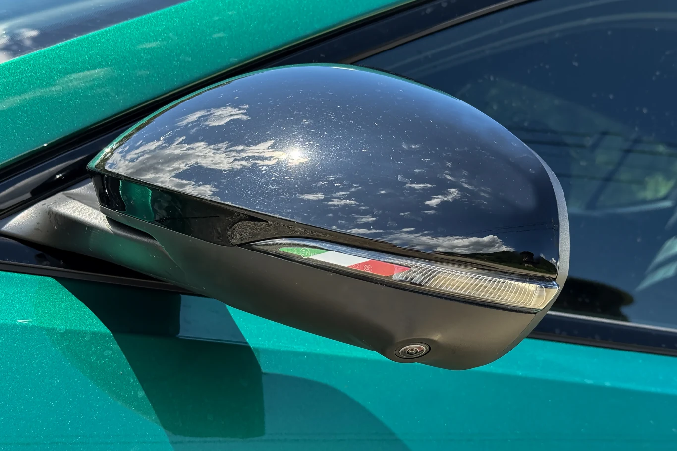 Czarne lusterko boczne samochodu w kolorze zielonym z odbiciem chmur na jego powierzchni, na lusterku widoczny delikatny akcent w barwach włoskiej flagi oraz kamera umieszczona u jego podstawy.