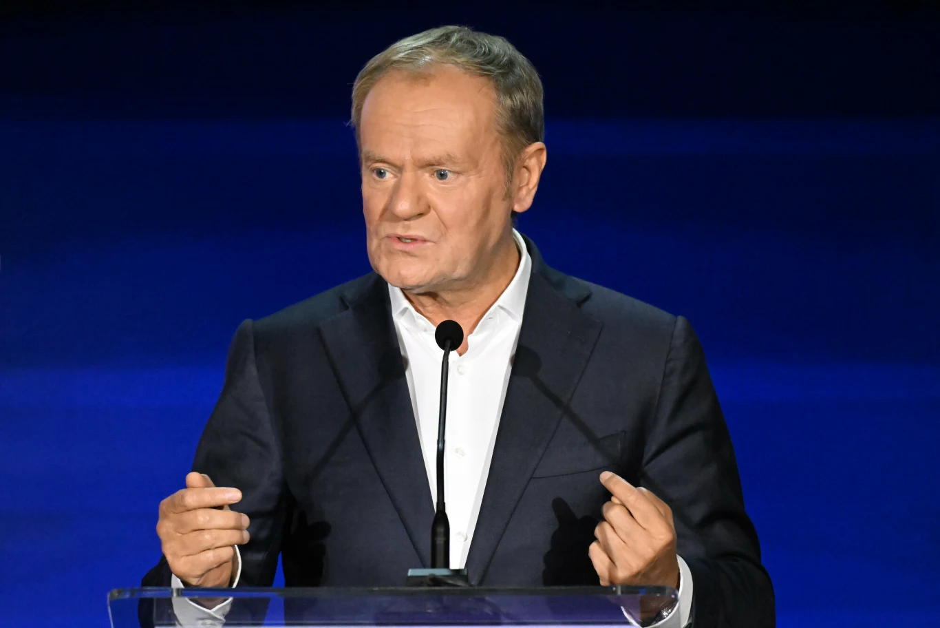 Premier Donald Tusk skomentował aferę wokół sprzedaży działki pod CPK Mężczyzna w garniturze stoi za przezroczystą mównicą, przemawia do mikrofonu na tle jednolitego niebieskiego tła.