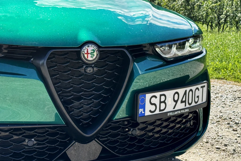 Scudetto, czyli trójkątna osłona chłodnicy to znak rozpoznawczy Alf od dziesięcioleci Zielony samochód marki Alfa Romeo z wyraźnie widoczną przednią atrapą chłodnicy, logo producenta oraz polską tablicą rejestracyjną na tle drogi i zieleni.