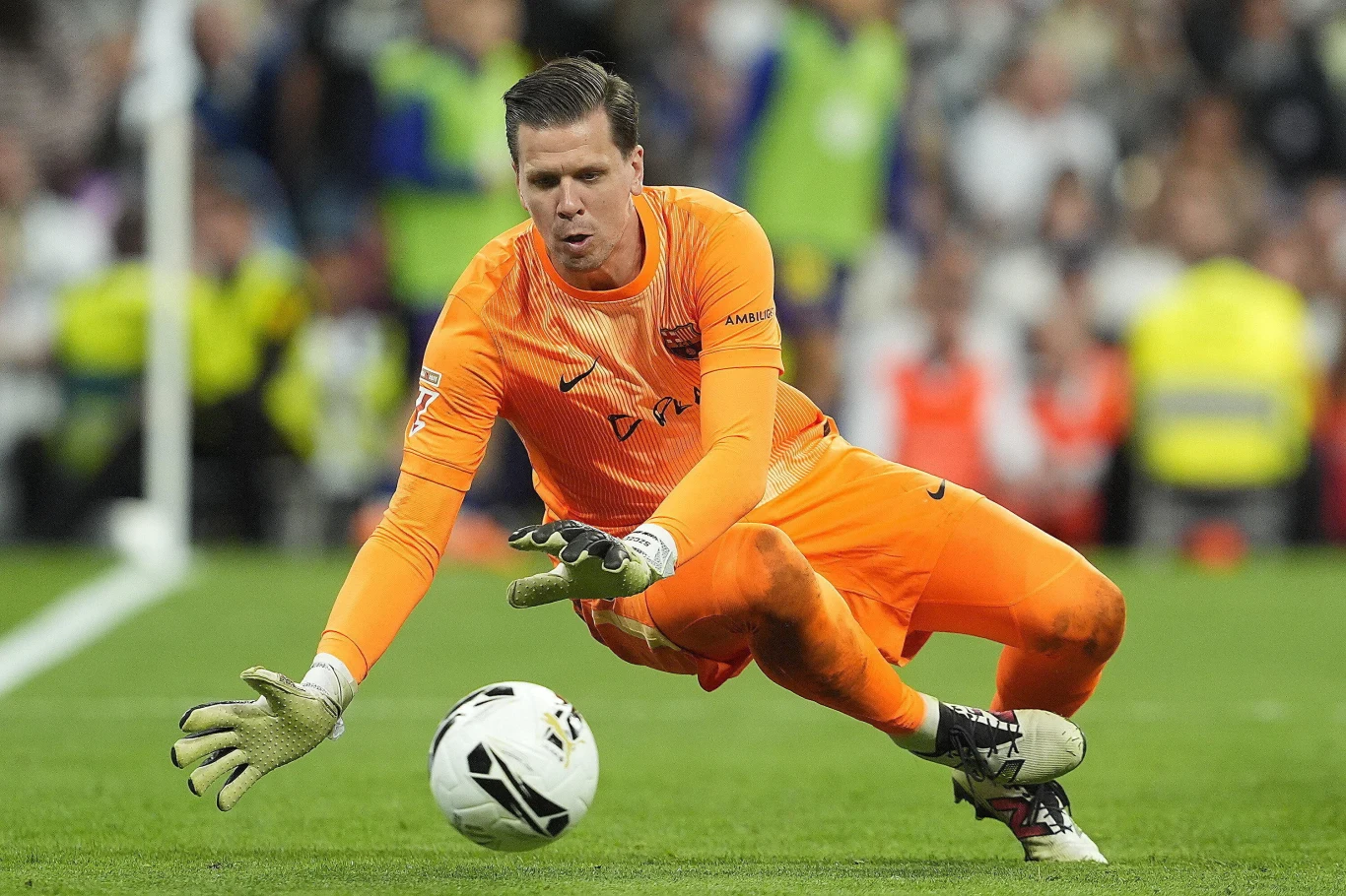 Wojciech Szczęsny w trakcie El Clasico Bramkarz w pomarańczowym stroju wykonuje interwencję podczas meczu piłkarskiego, skupiając się na łapaniu piłki, w tle widoczni kibice oraz fragment boiska.