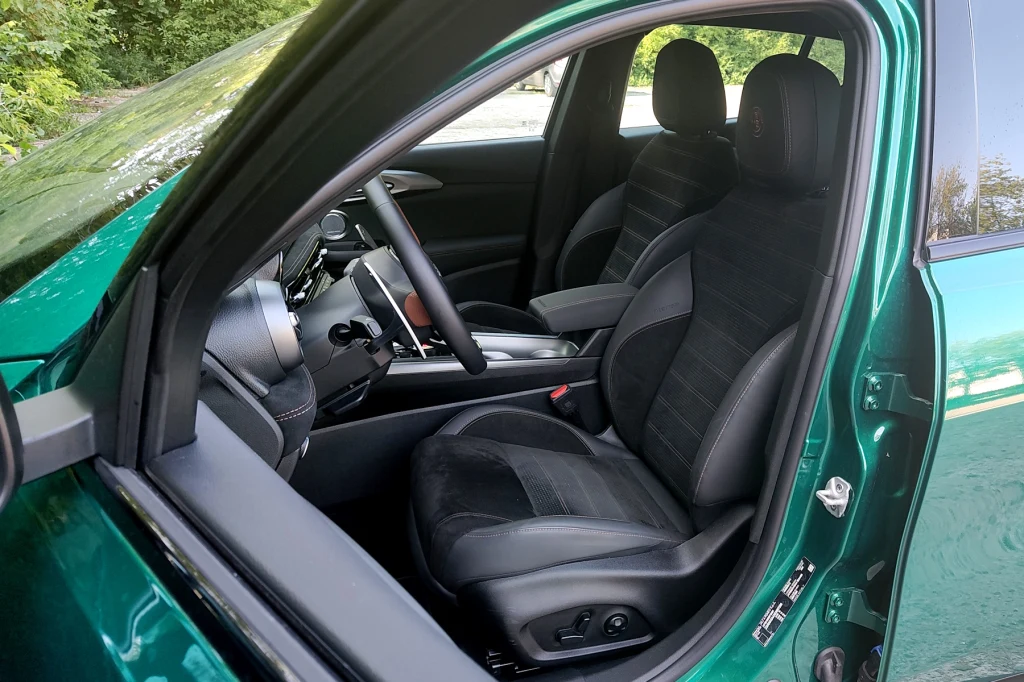 Fotele to mocna strona Alfy Romeo Tonale. Są wygodne, wszechstronnie regulowane i zapewniają dobre trzymanie Wnętrze nowoczesnego samochodu z widocznymi przednimi fotelami, deską rozdzielczą oraz kierownicą. Tapicerka czarna z eleganckimi przeszyciami, po lewej stronie wyraźnie widoczne drzwi kierowcy. Całość utrzymana w czystym, zadbanym stanie.
