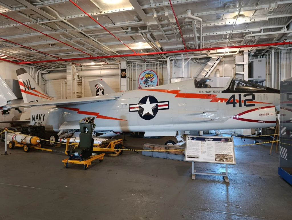 Myśliwiec F-8 Crusader na USS Hornet Amerykański odrzutowiec pokładowy F-8 Crusader o oznaczeniu NAVY 412, eksponowany w muzeum na pokładzie okrętu, otoczony barierkami ochronnymi i tablicą informacyjną, w tle widoczne elementy wyposażenia okrętowego.