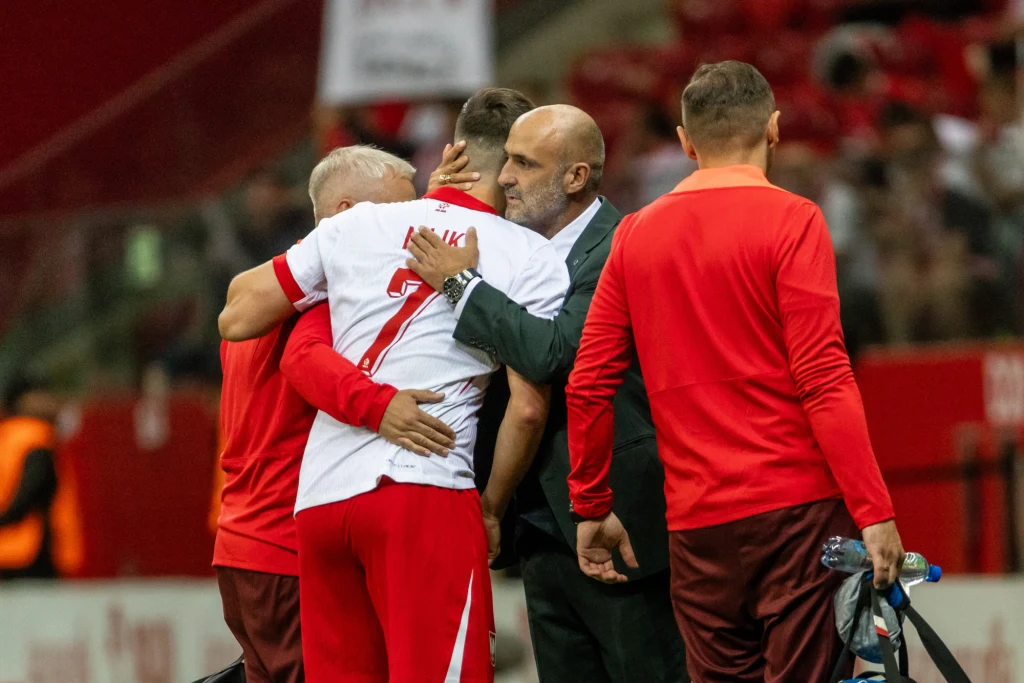 Arkadiusz Milik od momentu doznania kontuzji w sparingu z Ukrainą wciąż nie wrócił do gry Piłkarz ubrany w biało-czerwoną koszulkę z numerem 7 jest obejmowany przez starszego mężczyznę w garniturze na tle stadionu, wokół nich znajdują się inni członkowie drużyny lub sztabu szkoleniowego.