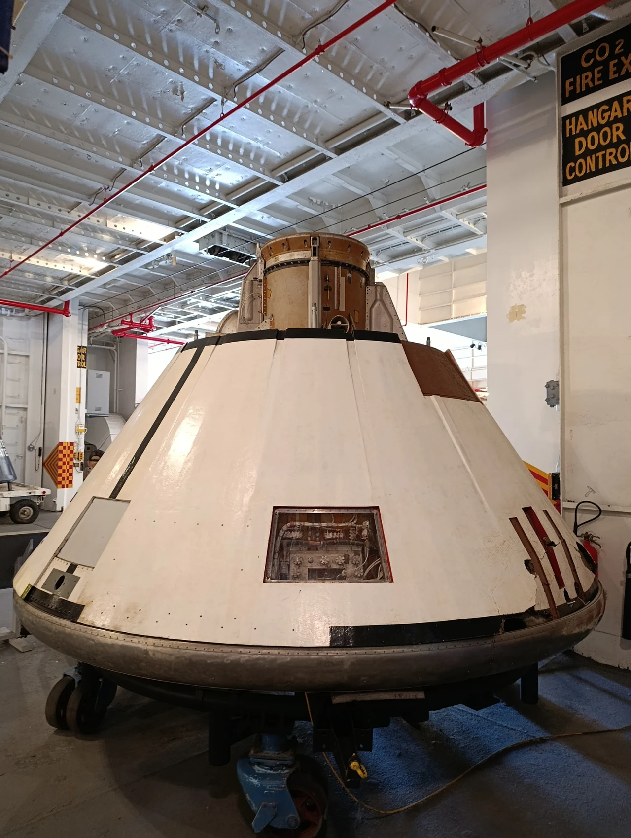 Kapsuła misji AS-202 na USS Hornet Duża kapsuła kosmiczna o stożkowatym kształcie, wykonana z białego metalu, umieszczona w hangarze z różnymi urządzeniami technicznymi i rurami widocznymi na suficie.