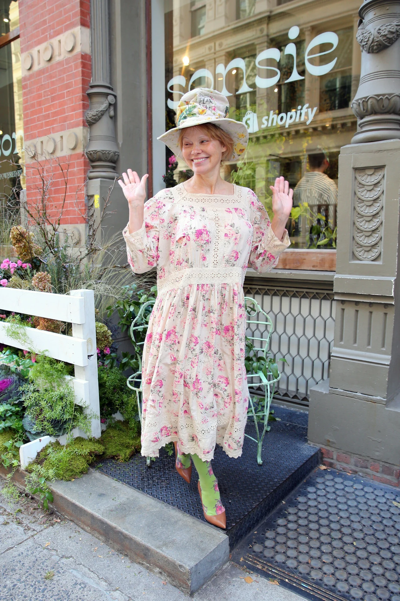 Pamela Anderson w oryginalnej, kwiatowej stylizacji Kobieta w sukience w kwiaty, uśmiechnięta, z uniesionymi rękami, pozuje przed wejściem do sklepu ozdobionego roślinami. Ma na głowie kapelusz z kwiatami, a tło stanowią witryna sklepu i miejskie otoczenie.