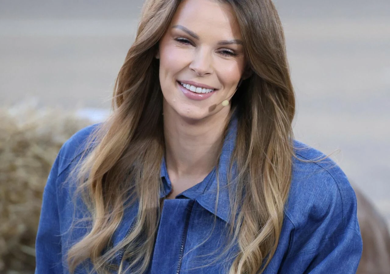 Małgorzata Tomaszewska Uśmiechnięta kobieta o długich, falowanych włosach w odcieniach brązu, ubrana w niebieską kurtkę, mikrofon przy twarzy, siedząca na tle rozmytego otoczenia z jasnym światłem.