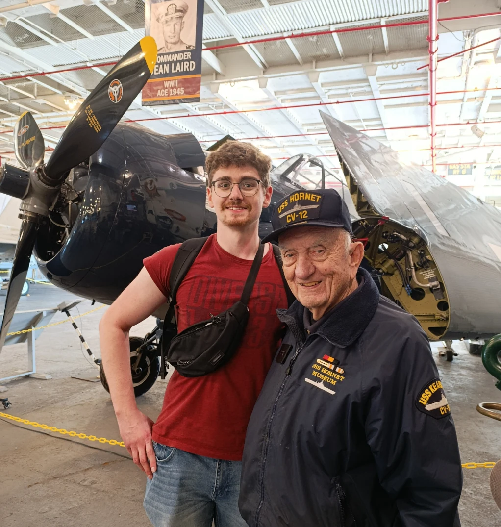 Część przewodników służyło na USS Hornet. Jednym z nich jest Paul Ackerman, który jest obecnie najstarszym weteranem, pracującym na lotniskowcu, na którym służył w latach 50. Dwóch mężczyzn pozuje przed historycznym samolotem w muzeum lotnictwa, starszy z nich ubrany w kurtkę i czapkę z napisem USS Hornet, drugi w czerwonej koszulce i okularach, obok widoczne tablice informacyjne.