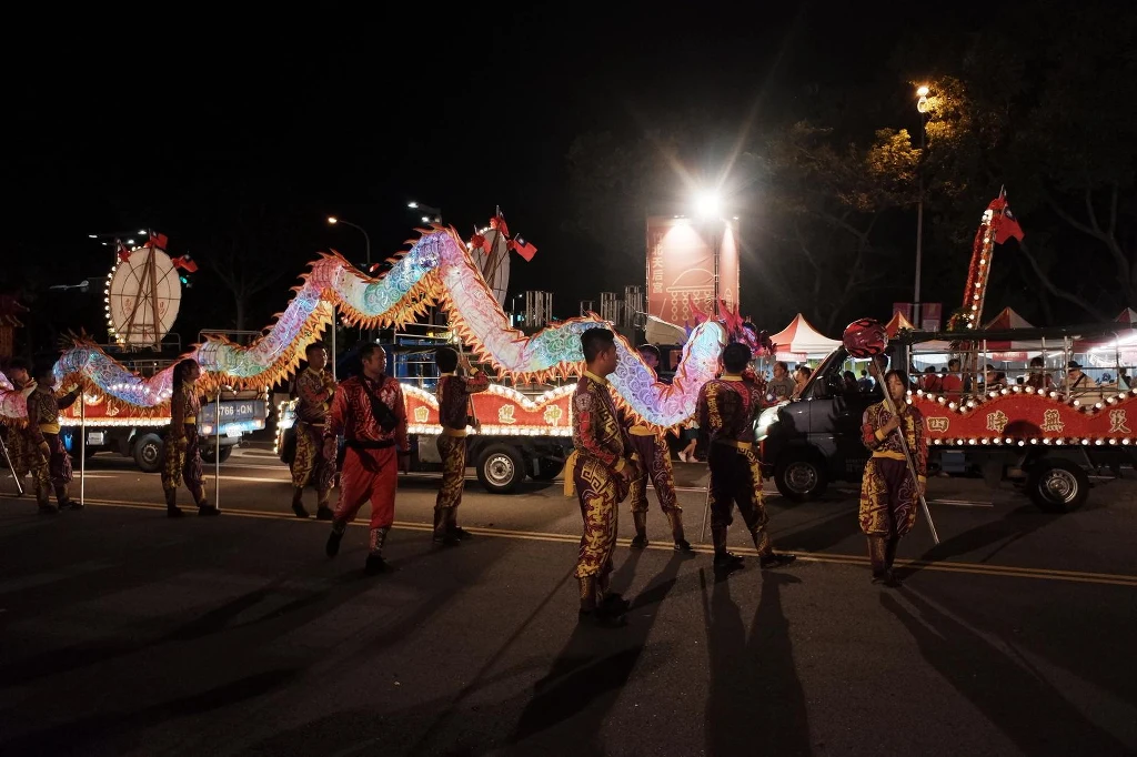 Wszystkich Świętych na Tajwanie. "Duchy wracają na ziemię, trzeba oddać im ofiarę" Kolorowy pochód nocą z tradycyjnym tańcem smoka, grupa osób ubrana w jasne stroje niesie długi, oświetlony smokowy kostium, w tle widoczne są ozdobione pojazdy oraz jasne światła.