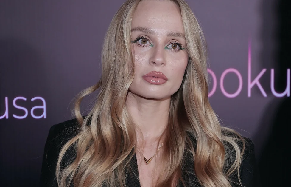 Sylwia Gaczorek Blondynka z długimi falowanymi włosami pozuje na tle ciemnego, rozmytego bannera z jasnymi napisami, ubrana w czarną marynarkę i delikatny naszyjnik, z wyrazistym makijażem podkreślającym oczy.