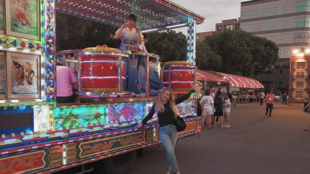 Kolorowa parada uliczna z dekoracyjnym, oświetlonym pojazdem, na którym kobieta gra na dużych bębnach. Przed pojazdem stoi kobieta pozująca do zdjęcia. W tle znajduje się kilka osób oraz namioty jarmarczne.