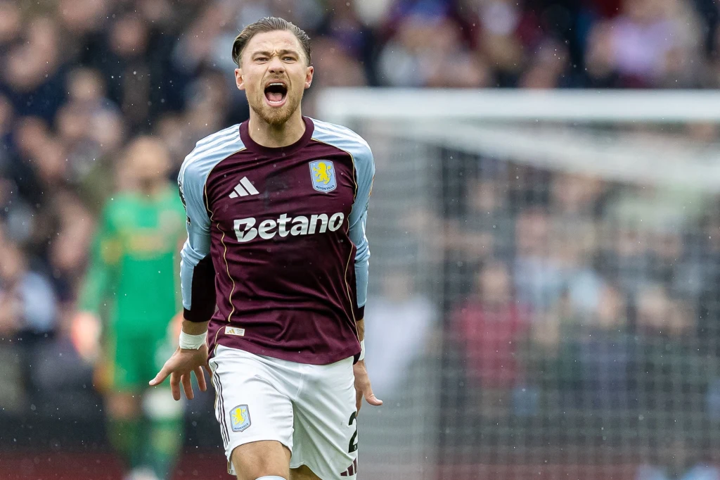 Piłkarz drużyny Aston Villa w bordowym stroju biegnie po boisku podczas meczu, wyrażając silne emocje krzykiem, w tle rozmyta publiczność i bramkarz w zielonym stroju.