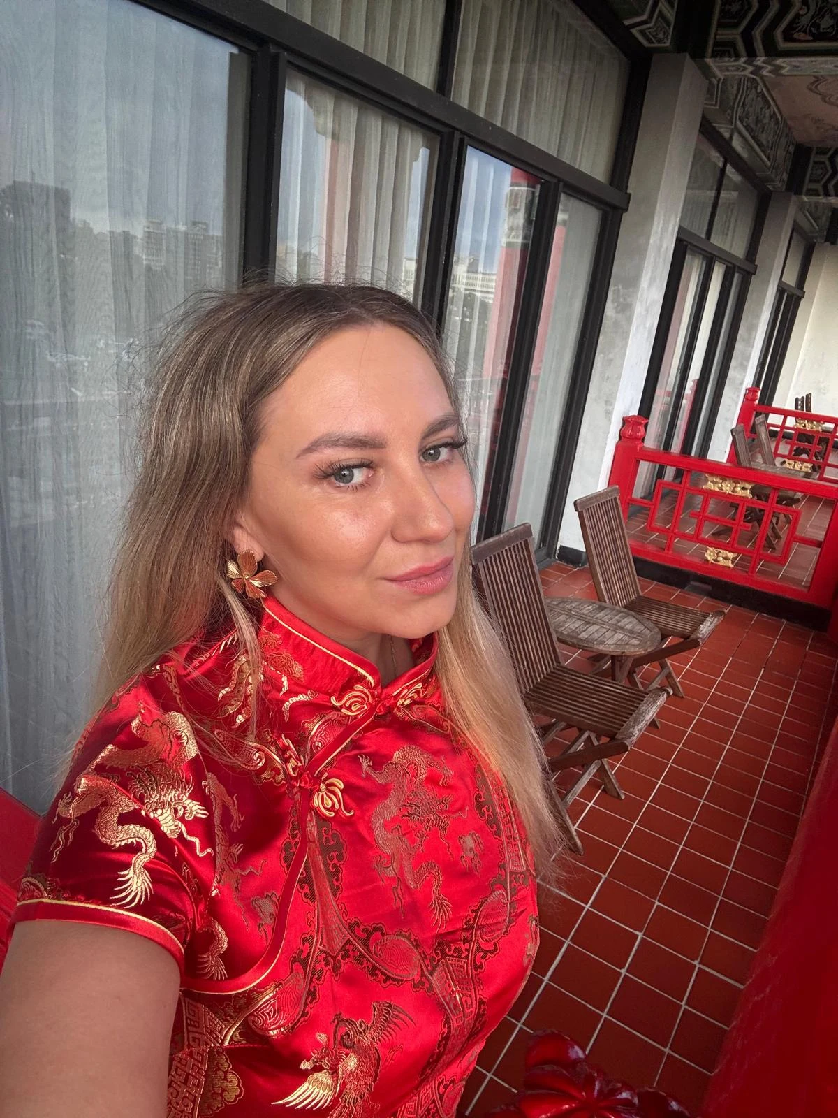 Wszystkich Świętych na Tajwanie. "Duchy wracają na ziemię, trzeba oddać im ofiarę" Kobieta w czerwonej, tradycyjnej chińskiej sukni z haftowanymi złotymi smokami stoi na tarasie z czerwonymi balustradami i drewnianymi krzesłami. W tle widoczne duże okna z zasłonami oraz fragment eleganckiego dachu.