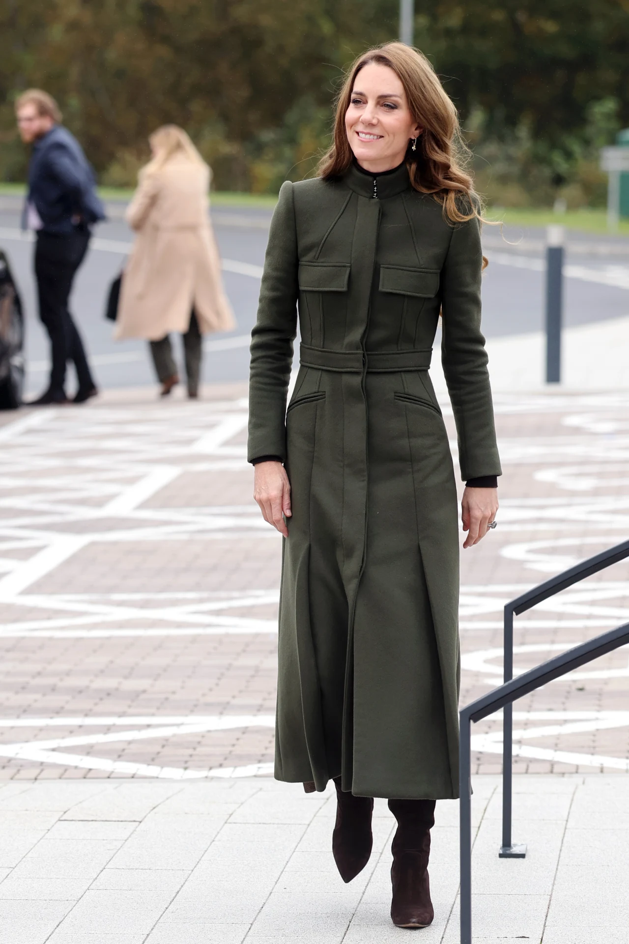 Księżna Kate uwielbia zielone płaszcze. Dodają jej szyku Elegancko ubrana kobieta w długim, zielonym płaszczu i ciemnych butach stoi na zewnątrz na chodniku, w tle widać dwie osoby idące oraz samochód zaparkowany przy ulicy, otoczenie miejskie z zielenią.