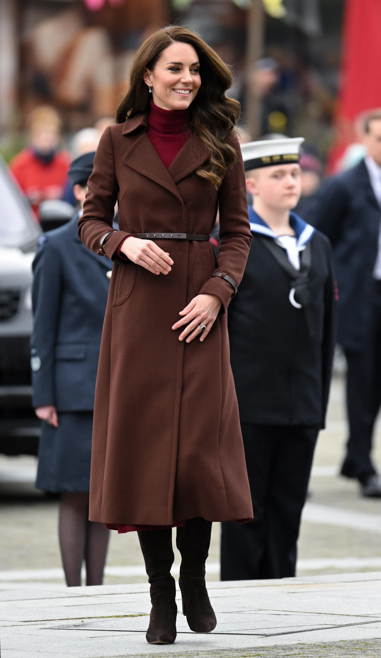 Brązowe zamszowe buty to częsty zimowy wybór księżnej Kate Elegancko ubrana kobieta w brązowym płaszczu i bordowym golfie stoi na zewnątrz, w tle widoczne są osoby w wojskowych mundurach oraz rozmyte sylwetki przechodniów.