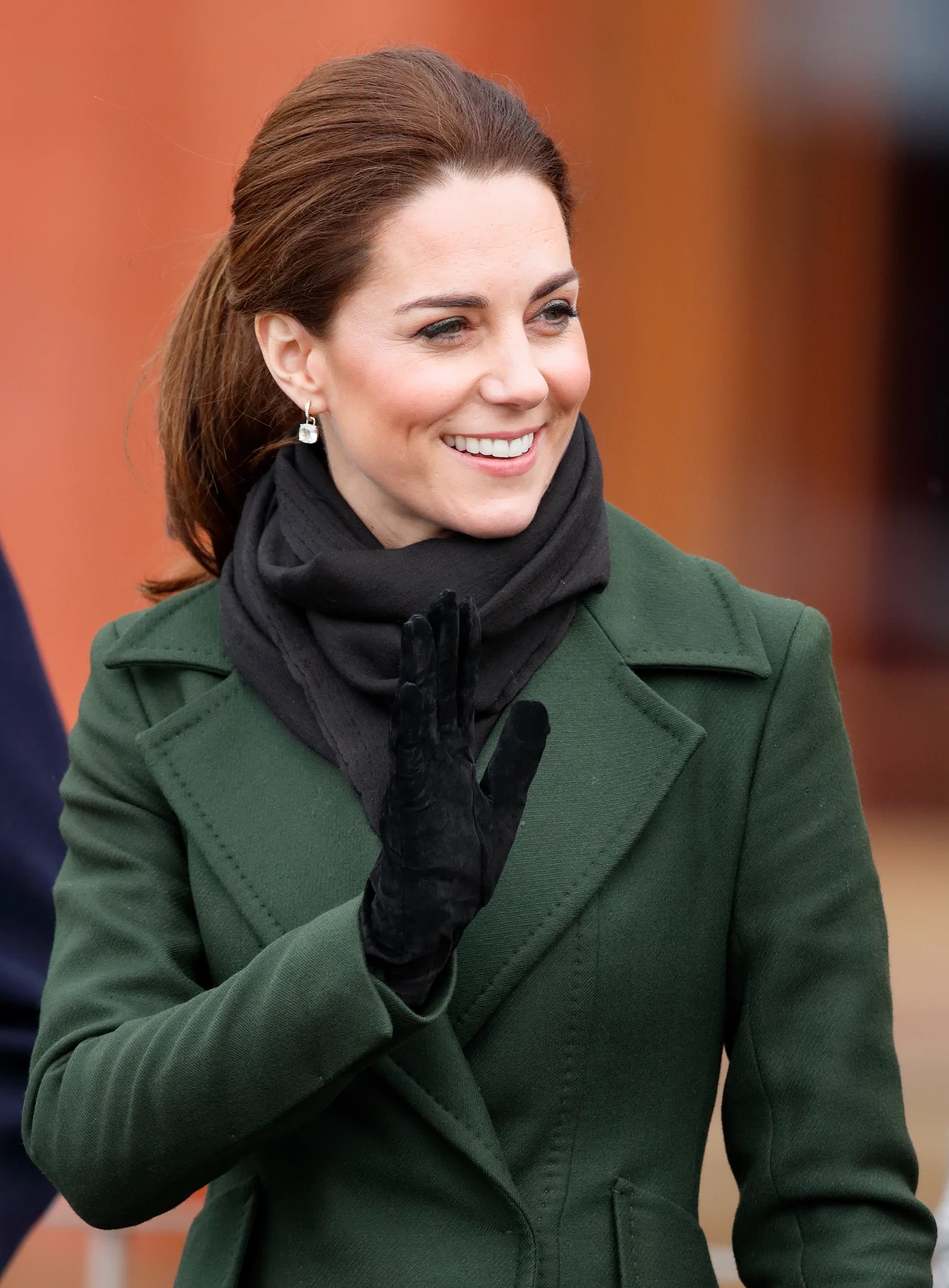 Księżna Kate uwielbia rękawiczki z wełny merino lub z zamszu Uśmiechnięta kobieta z upiętymi włosami ubrana w zielony płaszcz, czarny szalik i czarne rękawiczki, macha ręką w geście powitania.