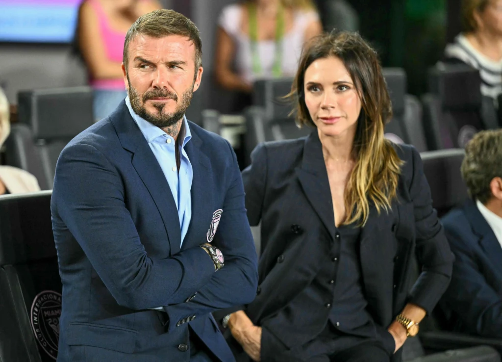 Victoria Beckham na meczu z Davidem Dwie elegancko ubrane osoby siedzą obok siebie na trybunach stadionu, mężczyzna ubrany w granatowy garnitur z zamkniętymi ramionami obok kobiety w czarnym stroju, oboje patrzą w stronę boiska, w tle widoczne są inne osoby.