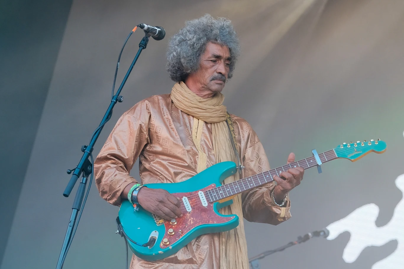 Ibrahim Ag Alhabib stoi na czele grupy Tinariwen Mężczyzna w tradycyjnym ubraniu z szalem gra na turkusowej gitarze elektrycznej na scenie, obok mikrofonu widoczny fragment tła koncertowego.