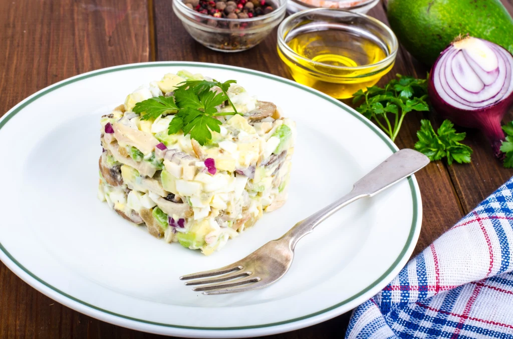 Sałatka pieczarkowa z dodatkiem jajek i ziemniaków jest nie tylko pożywna, ale i sycąca Porcja sałatki warstwowej na białym talerzu, ozdobiona listkiem natki pietruszki, ułożona estetycznie na kwadratową formę, obok widelca i ściereczki w kratkę, w tle świeża czerwona cebula, oliwa, przyprawy oraz awokado.