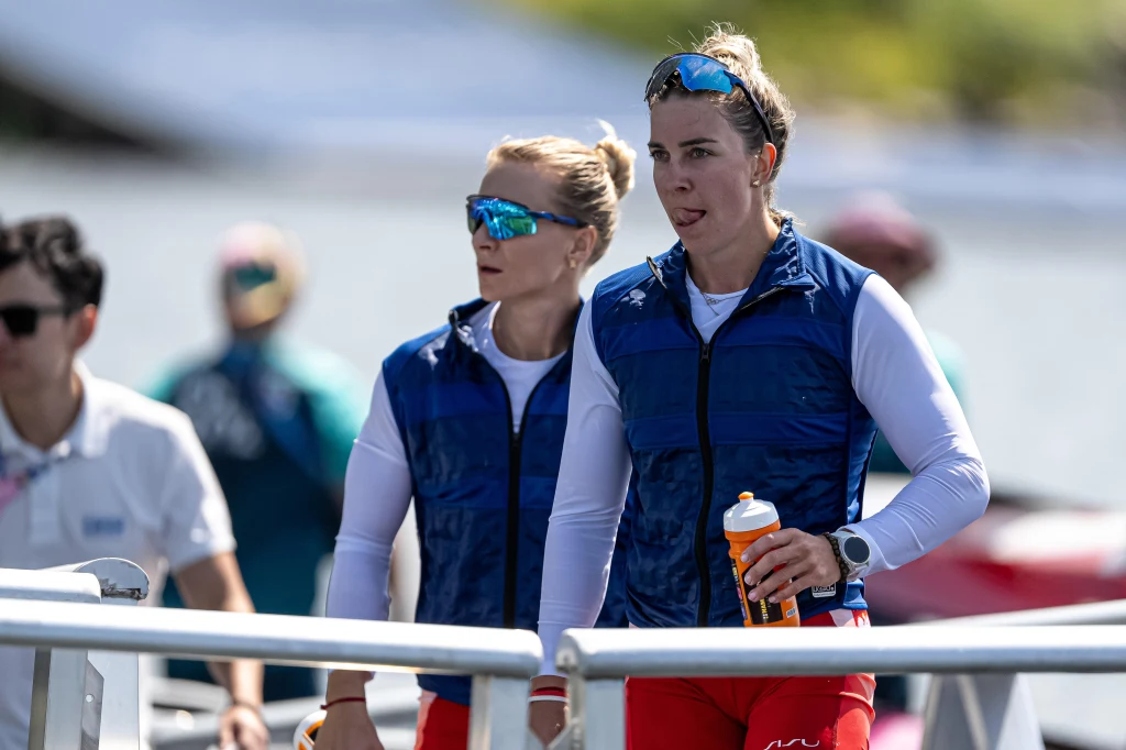 Anna Puławska (z prawej) i Karolina Naja Dwie zawodniczki w sportowych strojach i okularach przeciwsłonecznych stoją przy barierce na tle akwenu wodnego, jedna z nich trzyma bidon. W tle widoczne rozmazane postacie i łodzie.