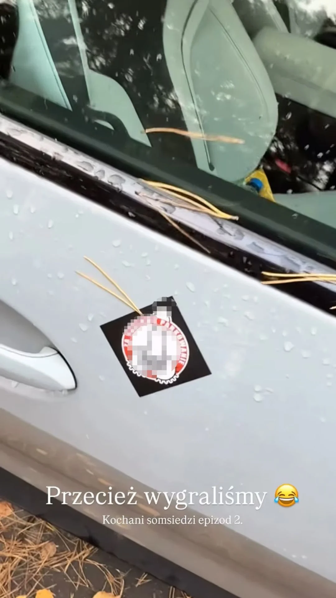 Fragment filmiku z Instagrama Karola Kłosa Białe auto zaparkowane na zewnątrz, na drzwiach znajduje się przyklejona kartka z zasłoniętym napisem oraz kilka rurek makaronu. Na zdjęciu widoczne są krople deszczu na karoserii i fragment wnętrza pojazdu.