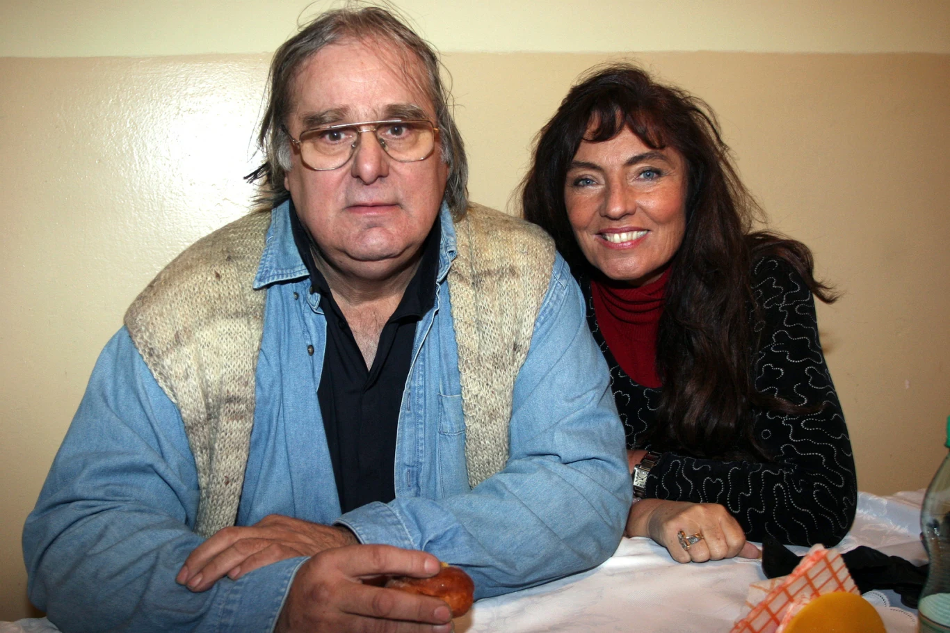 Marek Perepeczko i Agnieszka Fitkau-Perepeczko Mężczyzna w okularach oraz kobieta z długimi, ciemnymi włosami uśmiechają się, siedząc razem przy stole. Na stole widoczne owoce oraz butelka wody.