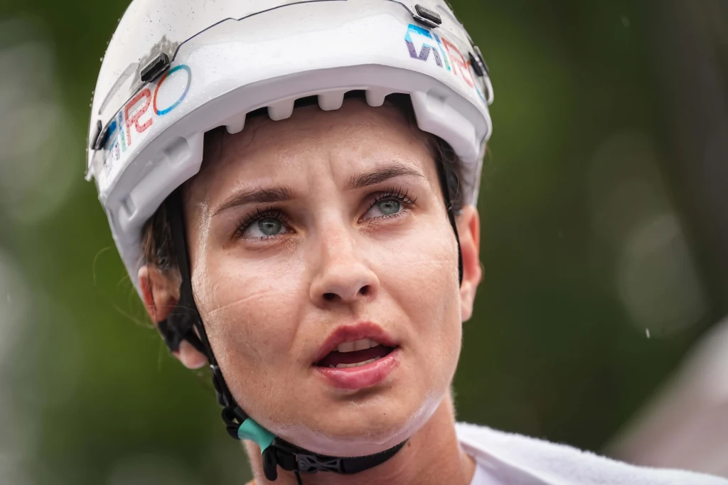 Młoda kobieta z jasnymi oczami i krótkimi włosami w białym kasku sportowym, lekko spocona, patrząca w dal z wyraźną determinacją na twarzy, w tle rozmyta zieleń.