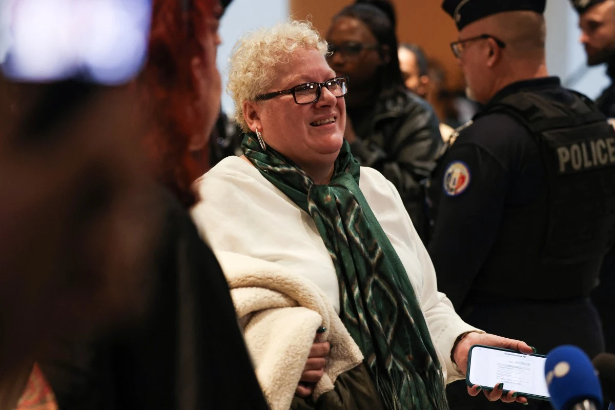 Uśmiechnięta kobieta z krótkimi, kręconymi włosami i okularami trzyma w ręce telefon oraz jasny płaszcz, wokół niej znajdują się osoby, w tym funkcjonariusze policji z widocznymi napisami na mundurach.