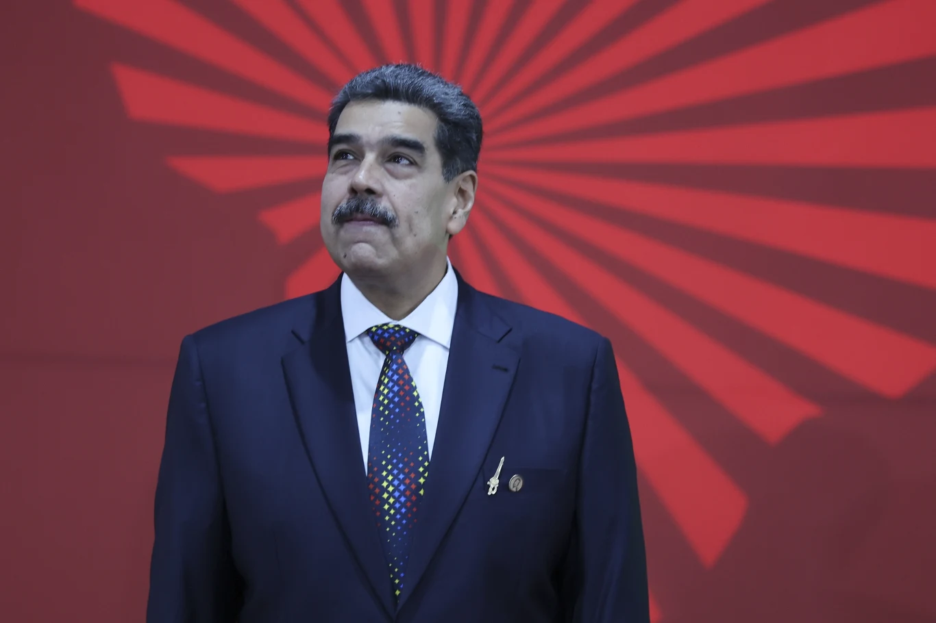 Nicolas Maduro Mężczyzna w garniturze i krawacie stoi na tle czerwonej ściany z geometrycznym wzorem przypominającym rozchodzące się promienie.