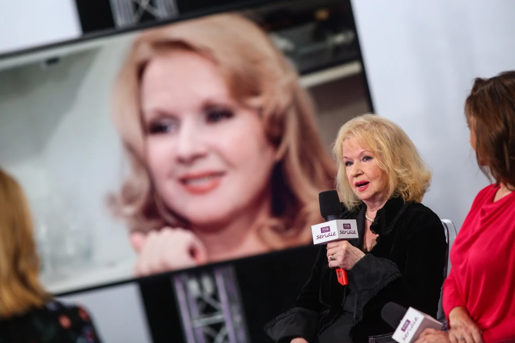 Dwie kobiety siedzące na scenie podczas wywiadu lub panelu dyskusyjnego, jedna z nich trzyma mikrofon i udziela odpowiedzi, w tle duży ekran z powiększonym wizerunkiem jednej z kobiet.