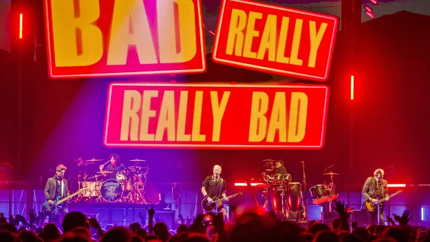 Koncert zespołu rockowego, na scenie pięciu muzyków grających na instrumentach w intensywnym, kolorowym oświetleniu. Nad sceną widoczne duże podświetlane napisy BAD, REALLY, REALLY BAD. Przed sceną duża grupa publiczności.