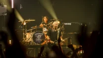 Energetyczna scena koncertowa z gitarzystą i perkusistą na tle jasnych reflektorów, tłum widzów unosi ręce w górę.