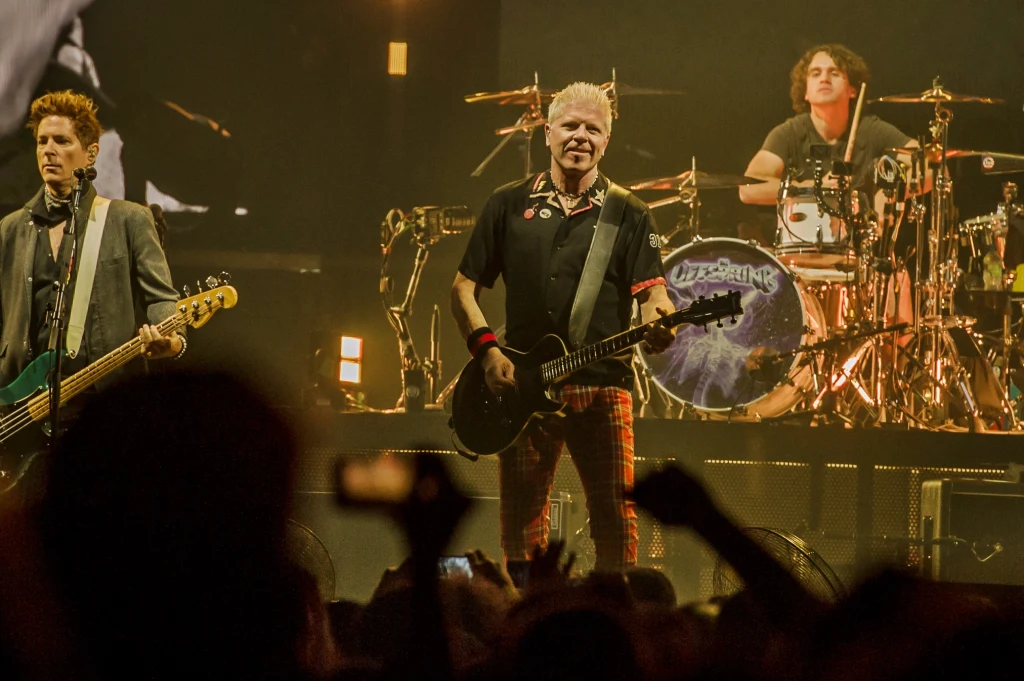 Offspring zagrali w Łodzi Trzech muzyków występuje na scenie, grając na gitarze basowej, elektrycznej i perkusji, otoczeni dynamicznym oświetleniem i tłumem publiczności pod sceną.