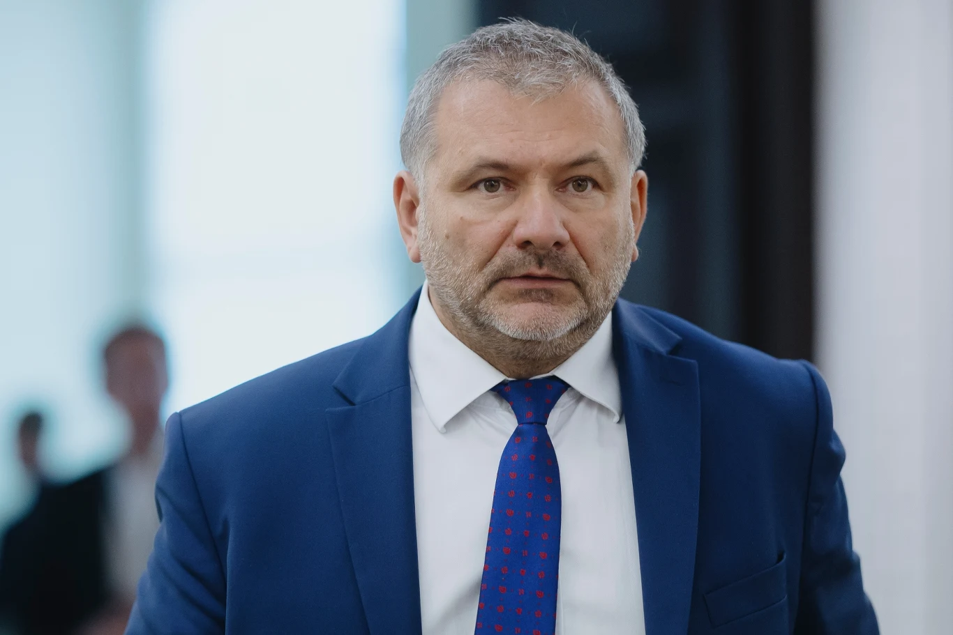 Waldemar Żurek zabrał głos w sprawie sprzedaży działki kluczowej dla CPK Mężczyzna w średnim wieku w granatowym garniturze i białej koszuli, z niebieskim krawatem w czerwone kropki, stoi w pomieszczeniu o jasnym tle. Ma krótką, siwiejącą brodę i poważny wyraz twarzy.