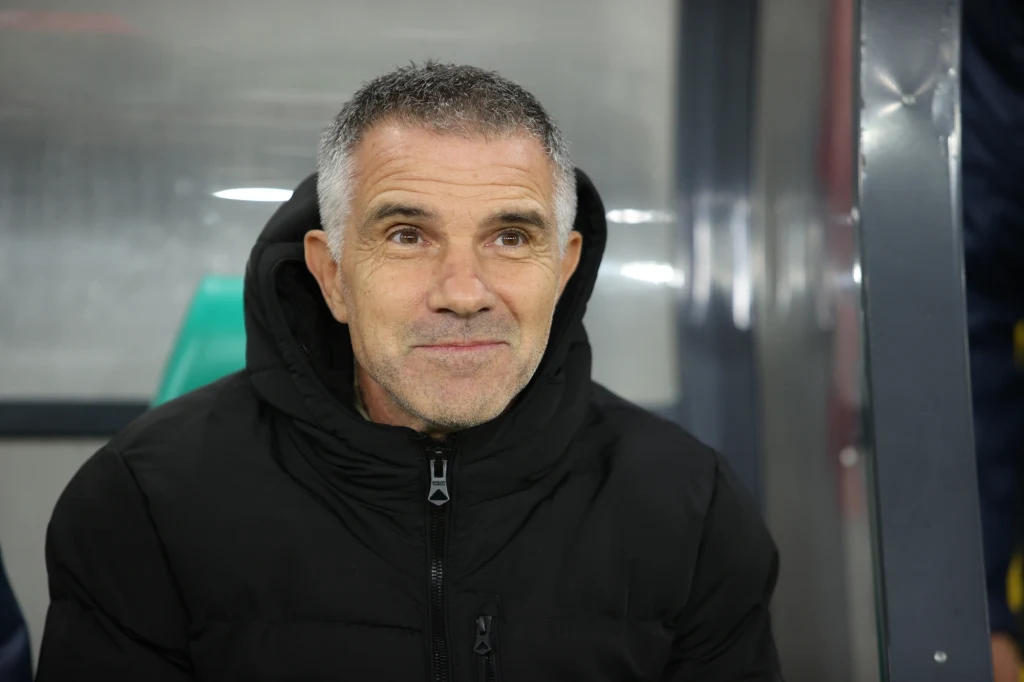 Trener krakowskiego zespołu, Gino Lettieri, przed pierwszym gwizdkiem był dobrej myśli Mężczyzna z krótkimi, siwiejącymi włosami i zarostem siedzi na ławce ubrany w czarną kurtkę, patrząc lekko w bok i delikatnie się uśmiechając.