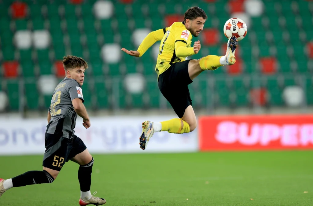 Piłkę w efektowny sposób przyjmuje Chuma. Tym razem nie pomógł jednak Wieczystej w odniesieniu zwycięstwa Dwóch piłkarzy podczas dynamicznej akcji na boisku, jeden z zawodników w żółto-czarnym stroju wykonuje wysoki podniebny kopnięcie piłki, drugi zawodnik w szarym stroju biegnie obok niego. W tle puste trybuny stadionu oraz rozmyte reklamy.
