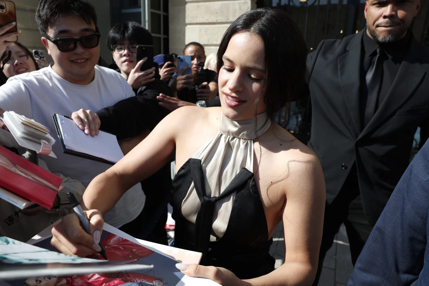 Rosalía nagrała teledysk w Polsce! Kobieta w eleganckiej sukni podpisuje autografy dla fanów, wokół niej zgromadzeni ludzie z aparatami i notesami, w tle widoczna ochrona i budynek.