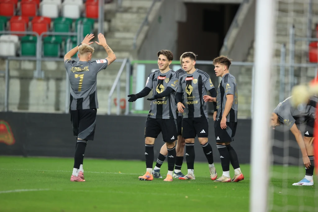 Pogoń Grodzisk Mazowiecki z kompletem punktów w starciu beniaminków z aspiracjami Grupa piłkarzy w szarych strojach wyraża radość po zdobyciu bramki, czterech zawodników stoi razem i gestykuluje, jeden z nich unosi ręce w geście triumfu, stadion w tle i widoczne trybuny.