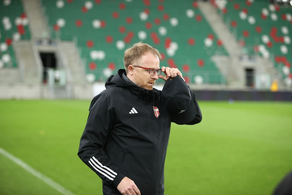 Piotr Stokowiec Mężczyzna w sportowej kurtce z herbem klubu piłkarskiego na piersi poprawia okulary na postawionym do połowy pustym stadionie.