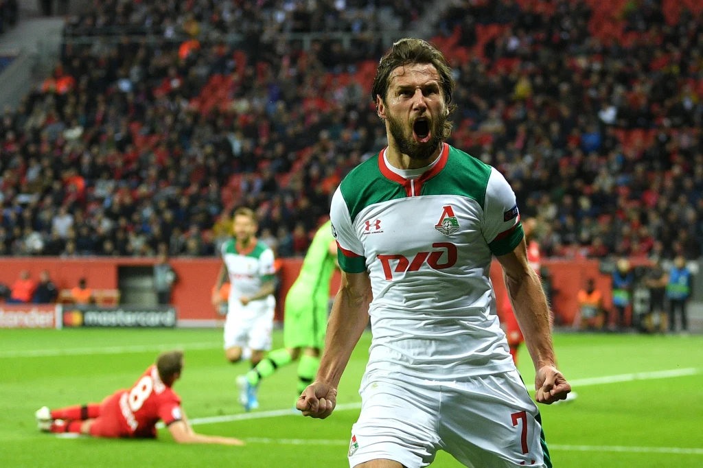 Krychowiak w barwach Lokomotiwu Moskwa Piłkarz w biało-zielono-czerwonym stroju drużyny Lokomotiv Moskwa wyraża intensywne emocje po zdobyciu gola, ściskając pięści i krzycząc. W tle widać kibiców na trybunach, a kilku innych zawodników znajduje się na murawie, jeden z nich leży na ziemi.