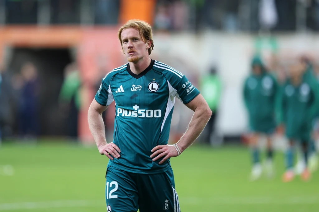 Radovan Pankov Piłkarz w stroju Legii Warszawa stoi na boisku z rękami opartymi na biodrach, wyraża zmęczenie lub rozczarowanie, w tle zamazane sylwetki innych zawodników i kibiców.