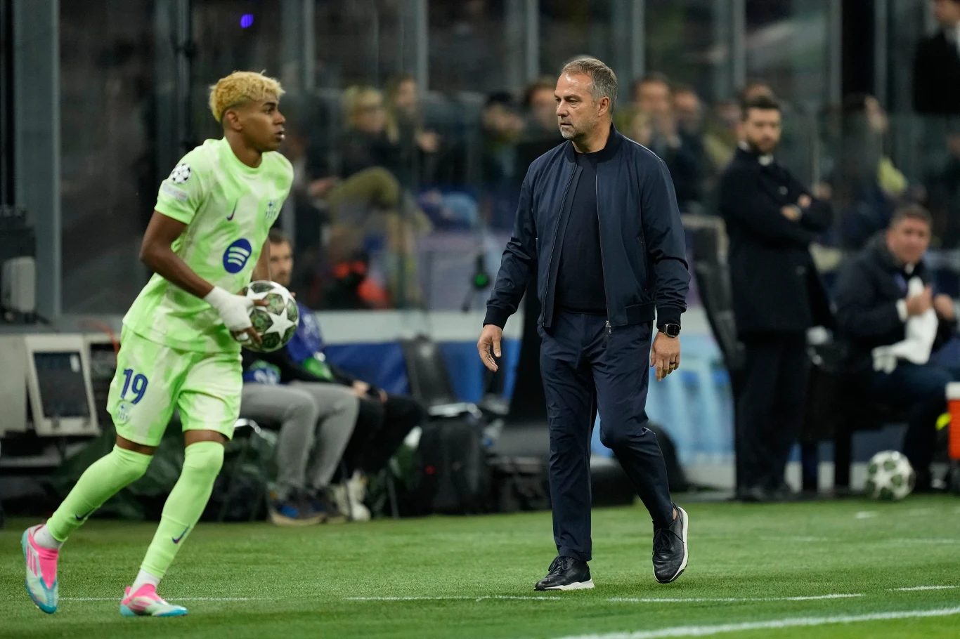 Zawodnik piłkarski w jasnozielonym stroju trzyma piłkę, prawdopodobnie przygotowując się do wyrzutu z autu, podczas gdy trener w ciemnej kurtce uważnie obserwuje boisko. W tle widoczni sędziowie i inni członkowie zespołu na ławkach rezerwowych.