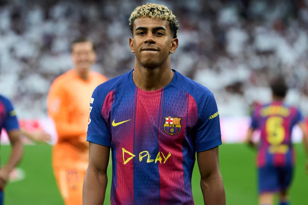 Piłkarz w granatowo-bordowej koszulce FC Barcelony stoi na boisku, w tle inni gracze oraz rozmazana trybuna stadionu.