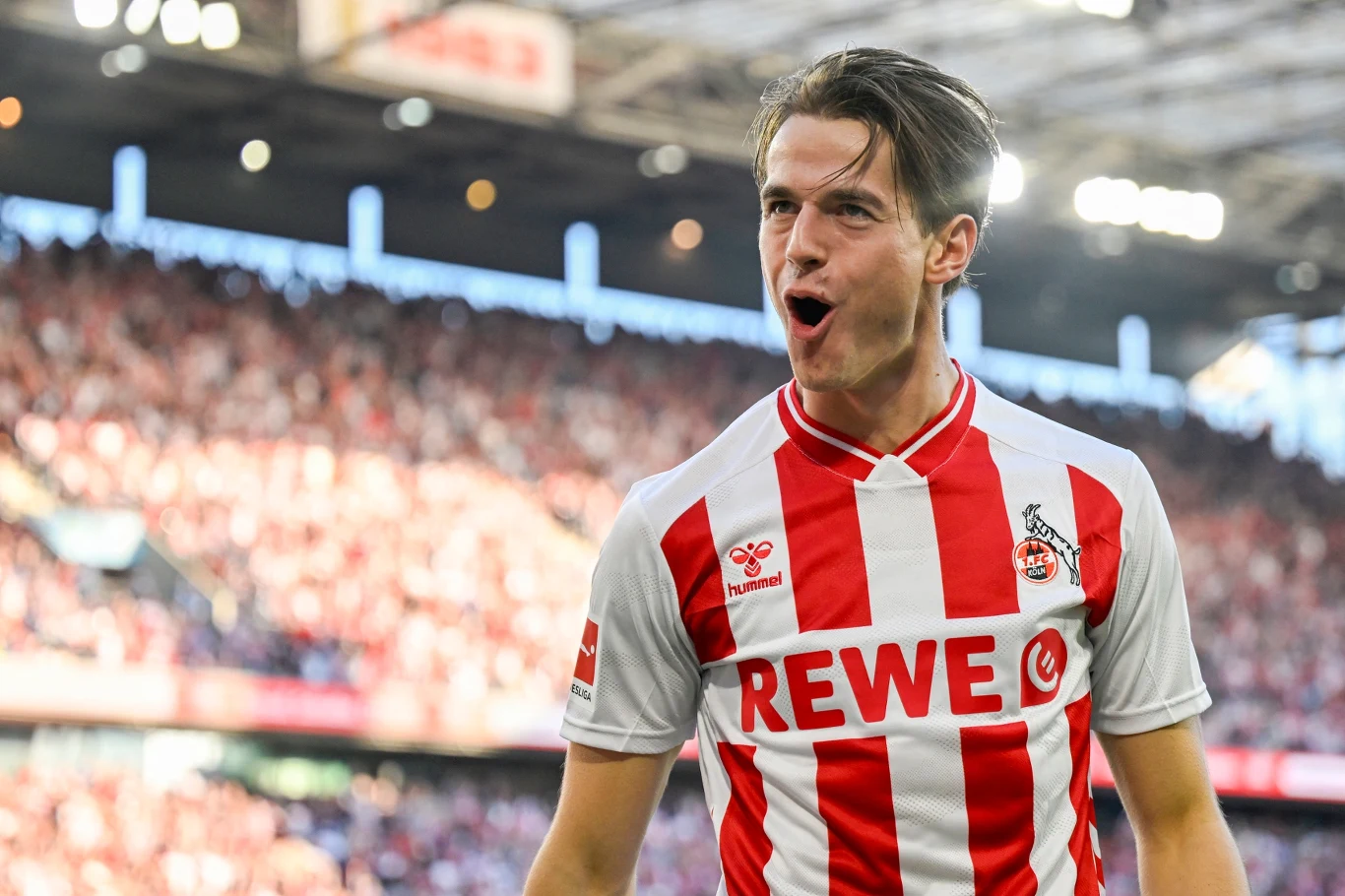 Jakub Kamiński Piłkarz w biało-czerwonej koszulce drużyny z herbem koziego kozła i napisem REWE na piersi, wyraża radość na tle pełnych trybun stadionu podczas meczu.