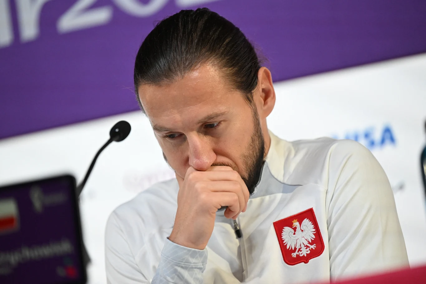 Mężczyzna w białej bluzie z godłem Polski na piersi, siedzący przy mikrofonie, pochylony i zamyślony, z ręką opartą o usta na tle fioletowego baneru.