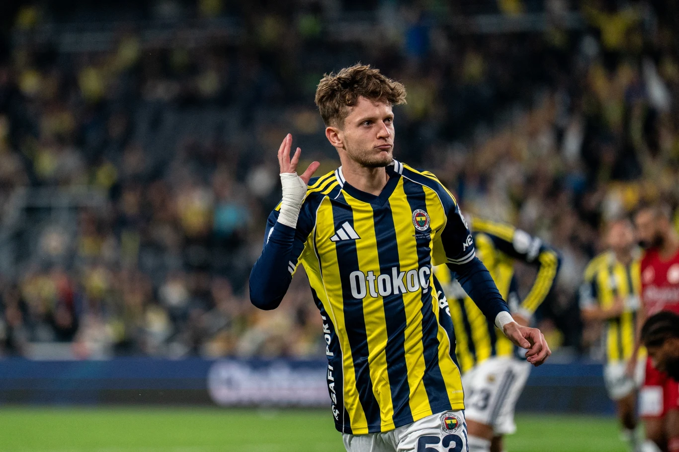 Piłkarz w żółto-granatowym stroju świętuje zdobycie gola na tle rozmytego tłumu kibiców oraz innych zawodników na boisku.