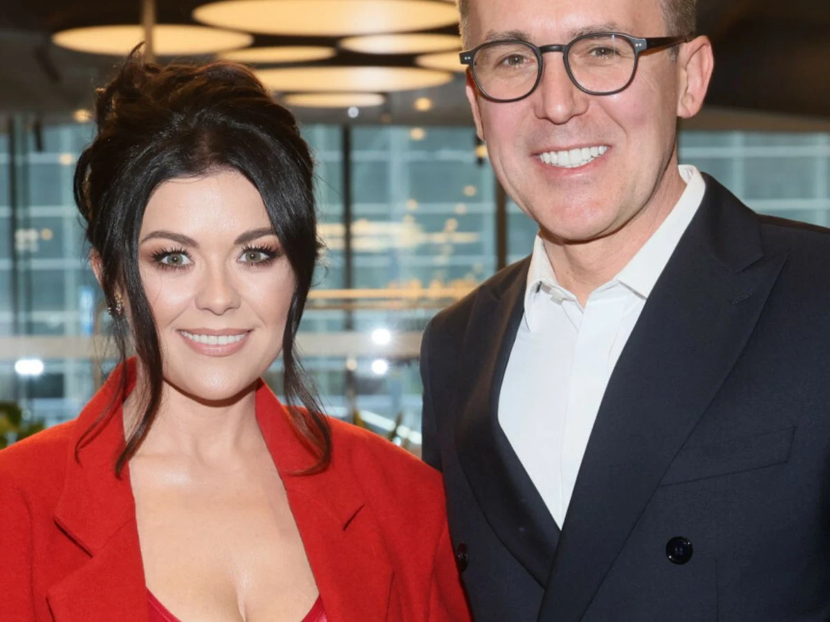 Katarzyna Cichopek i Maciej Kurzajewski Kobieta w czerwonym żakiecie i mężczyzna w garniturze stoją obok siebie, oboje uśmiechnięci, w tle nowoczesne wnętrze z dużymi oknami i widokiem na miejskie budynki.