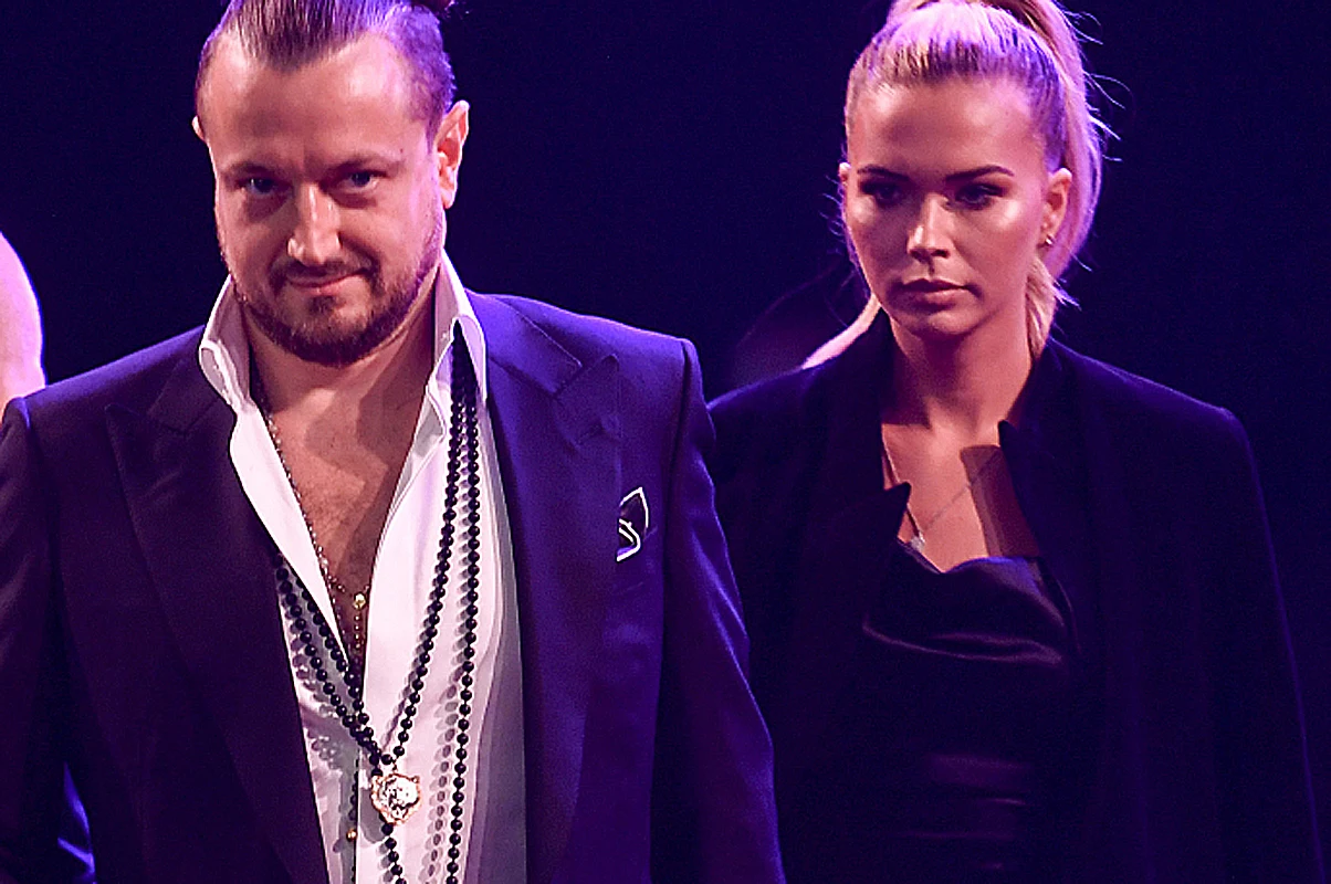 Baron i Kubicka Mężczyzna w eleganckim garniturze z rozpiętą białą koszulą, na szyi zawieszony duży naszyjnik. Obok stoi kobieta w czarnej stylizacji, włosy związane w kok, oboje w poważnych wyrazach twarzy, oświetleni fioletowym światłem.